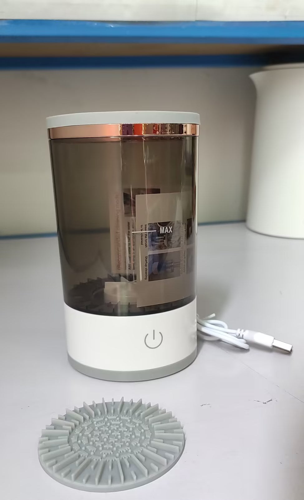 Clear cylindrical container with white base and rose gold accents on a gray surface.