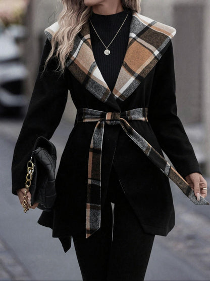Person wearing a black coat with plaid accents and a belt, holding a handbag.