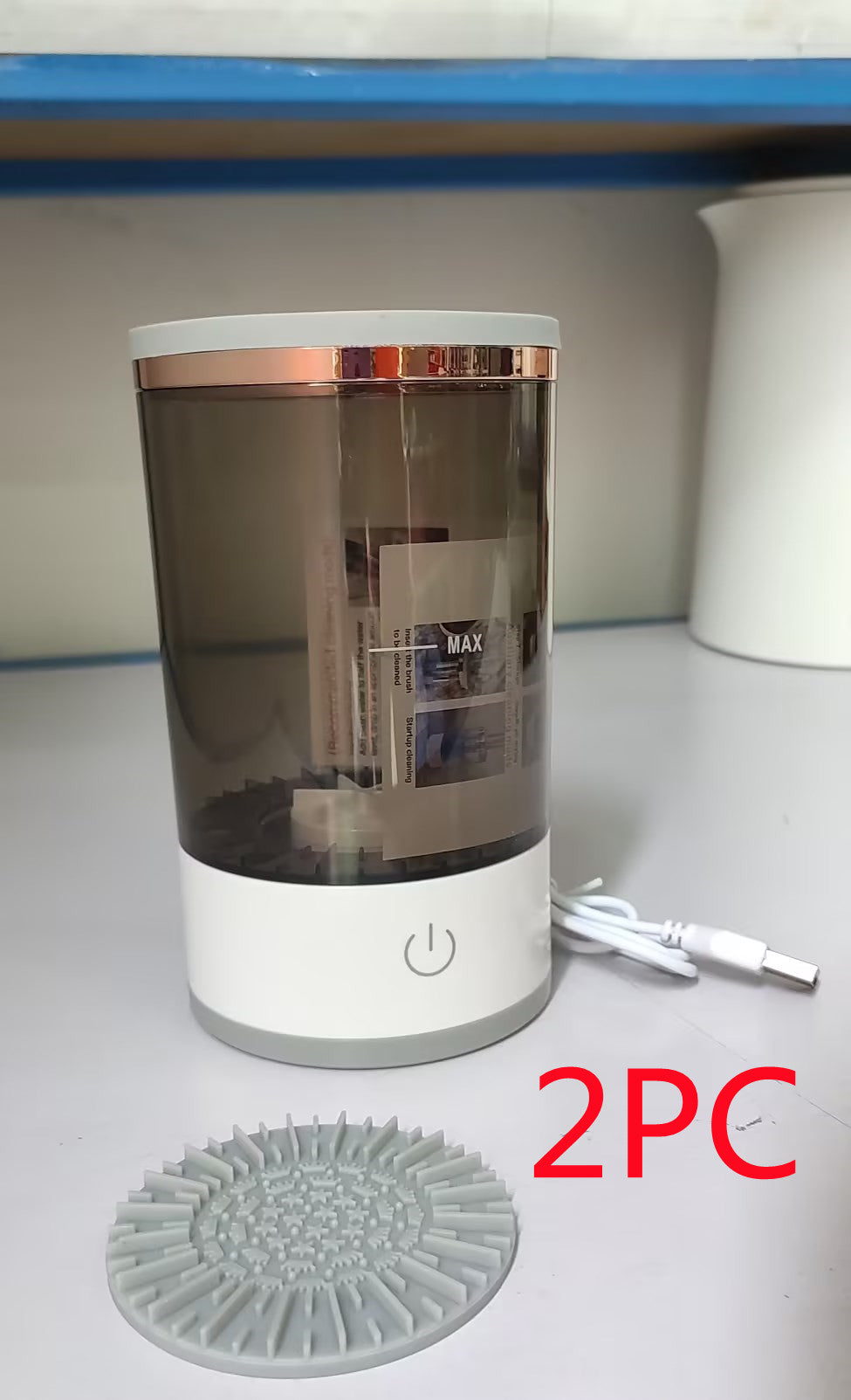 Transparent cylindrical container with rose gold accents on a white surface, accompanied by a gray silicone mat and white cable.